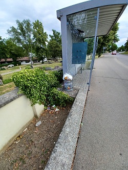Das Bild zeigt eine Bushaltestelle vor einem Friedhof, in deren Mülleimer eine Tüte mit Hausmüll steckt.