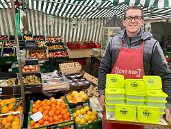 Auf dem Bild ist ein Mann zu sehen, der gelbe Frischhalteboxen in den Händen hält. Im Hintergrund liegt Obst und Gemüse, das an einem Marktstand angeboten wird.