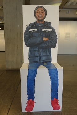 Das Foto zeigt ein Bild des Künstlers Jan-Hendrik Pelz. Das Bild zeigt einen Mann mit dunkler Hautfarbe, seine Arme sind vor der Brust verschränkt, er trägt eine Winterjacke und eine Mütze