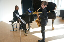 Auf dem Bild sind zwei Kinder zu sehen, die auf einem Flügel und einem Saxophon Musik machen.