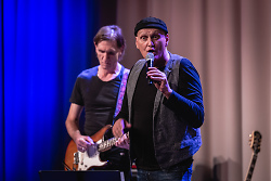 Das Bild zeigt Michael Flechsler mit einem Mikrophon in der Hand. Im Hintergrund spielt ein Gitarrist. 