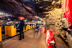 Auf dem Foto ist das Innere des Besucherbergwerks Tiefer Stollen zu sehen. Abgebildet ist der Zug, mit dem man ins Bergwerk einfährt. Im Zug sitzen Menschen.